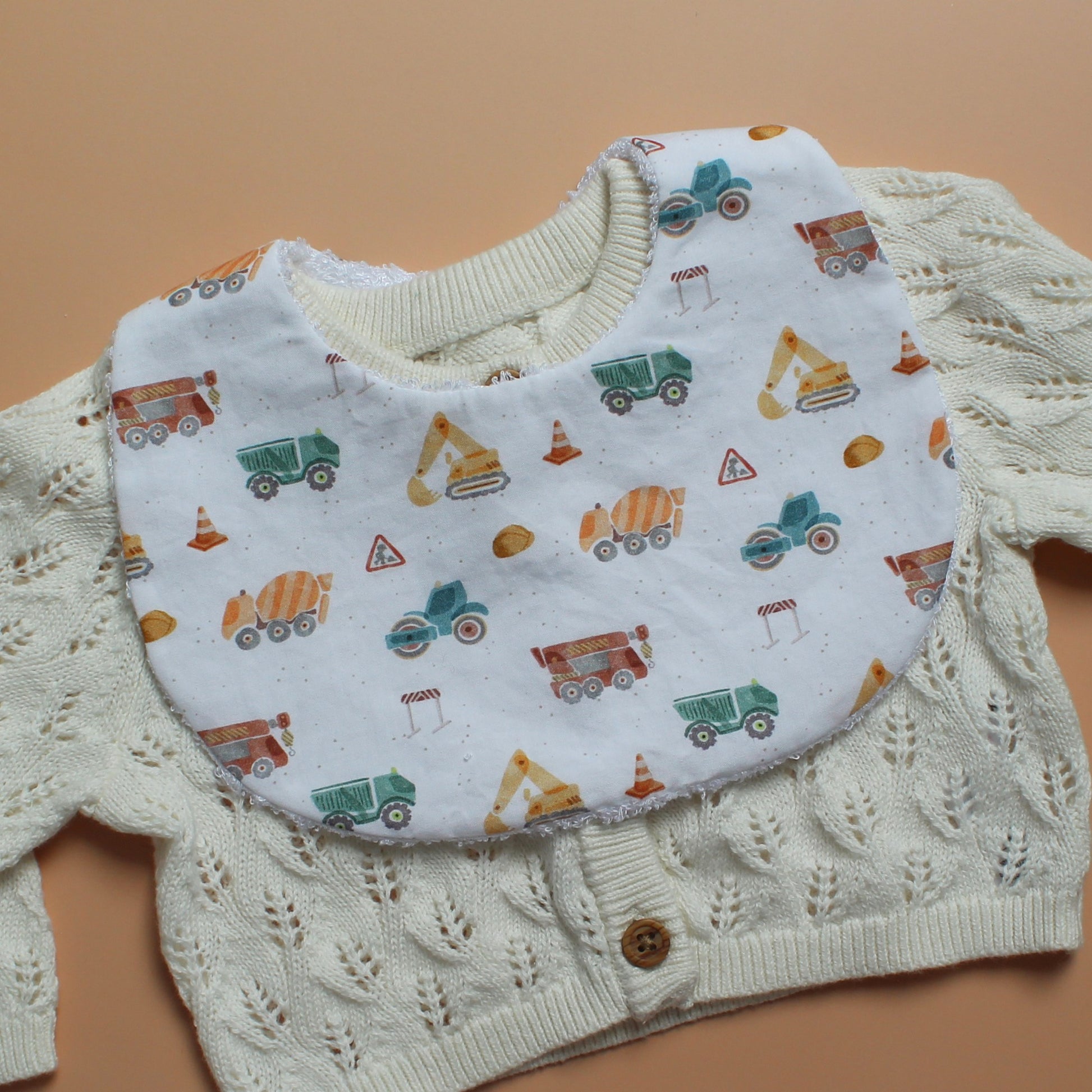 A baby bib with a construction vehicle print, layered over a cream-colored cable-knit cardigan sweater, laid flat on a solid tan background.