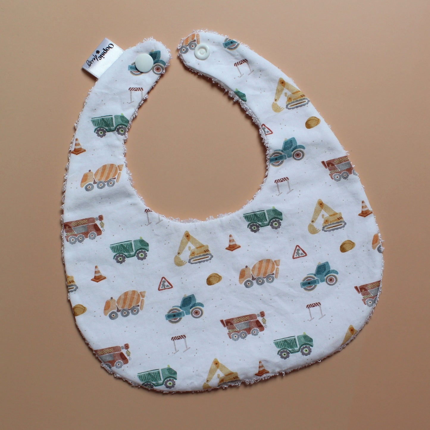 A white fabric baby bib with a colourful construction vehicle print, featuring snap closures at the top, laid flat on a solid tan background.