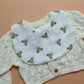 A white baby bib with a repeating pattern of small brown teddy bears and subtle gingham pattern, layered over a cream-coloured cable-knit baby cardigan sweater on a solid peach background.