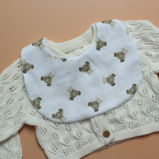 A white baby bib with a repeating pattern of small brown teddy bears and subtle gingham pattern, layered over a cream-coloured cable-knit baby cardigan sweater on a solid peach background.