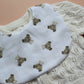 A close-up of a white fabric bib with a repeating teddy bear print and a subtle gingham pattern, laid over a cream-coloured cable-knit cardigan.