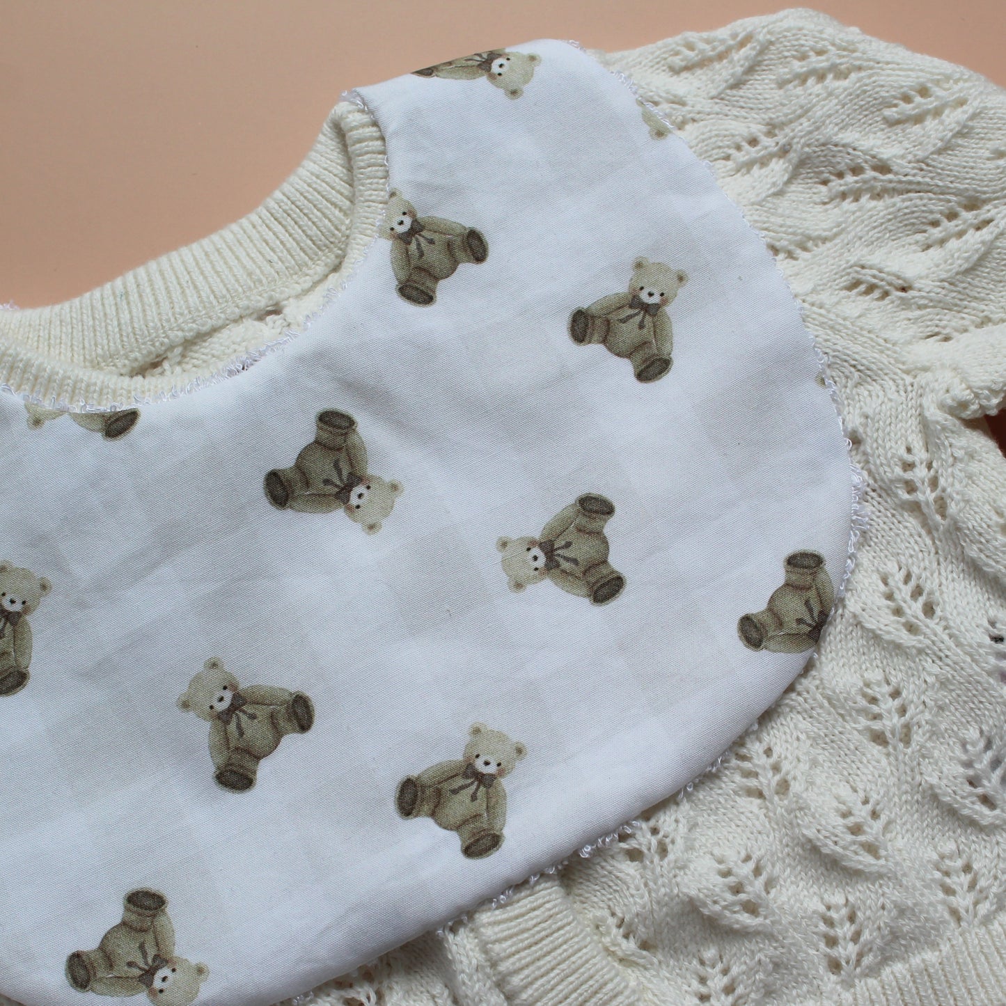 A close-up of a white fabric bib with a repeating teddy bear print and a subtle gingham pattern, laid over a cream-coloured cable-knit cardigan.