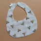 A white fabric baby bib with a repeating pattern of small brown teddy bears and subtle gingham pattern, featuring white snap closures at the top, laid flat on a solid peach-colored background.