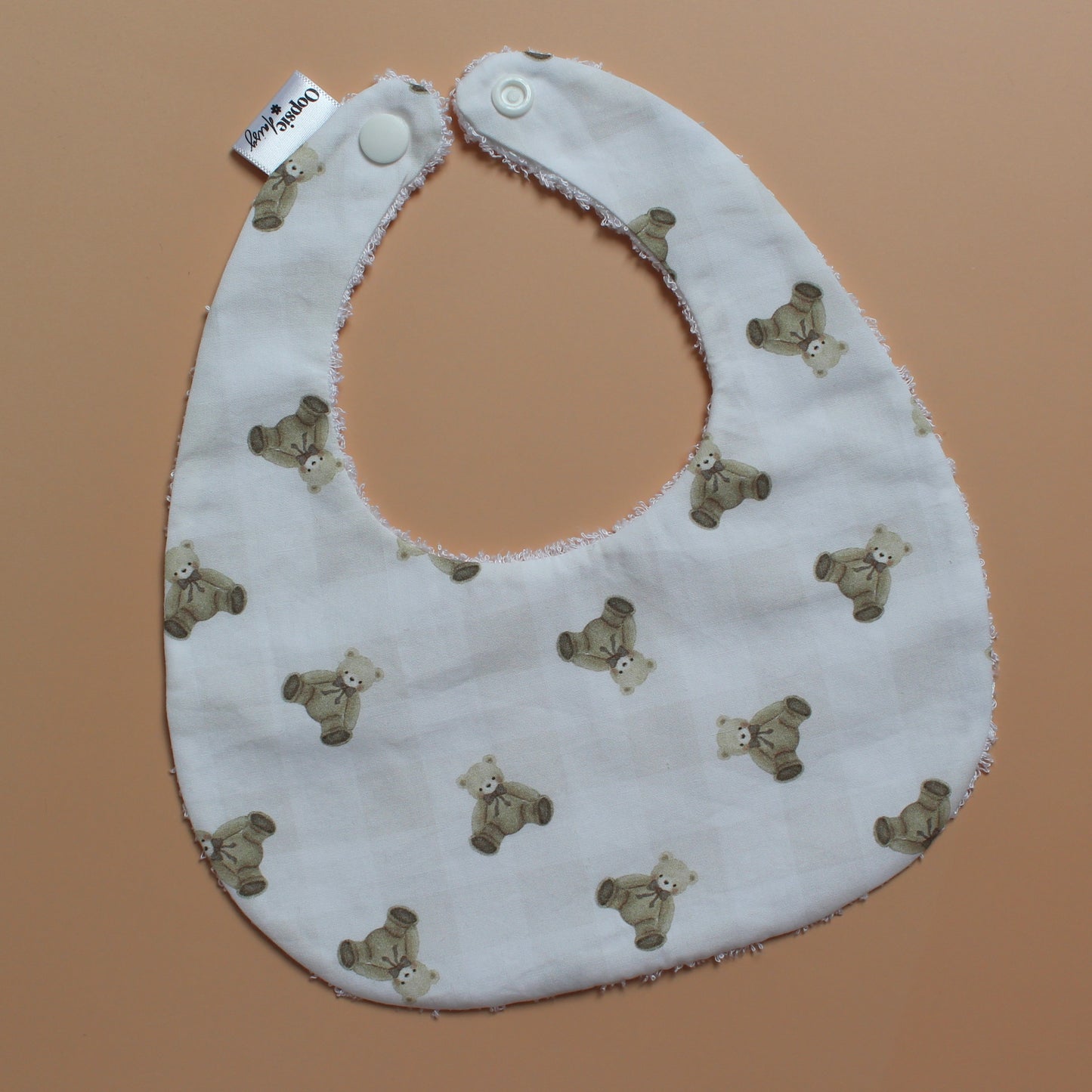 A white fabric baby bib with a repeating pattern of small brown teddy bears and subtle gingham pattern, featuring white snap closures at the top, laid flat on a solid peach-colored background.