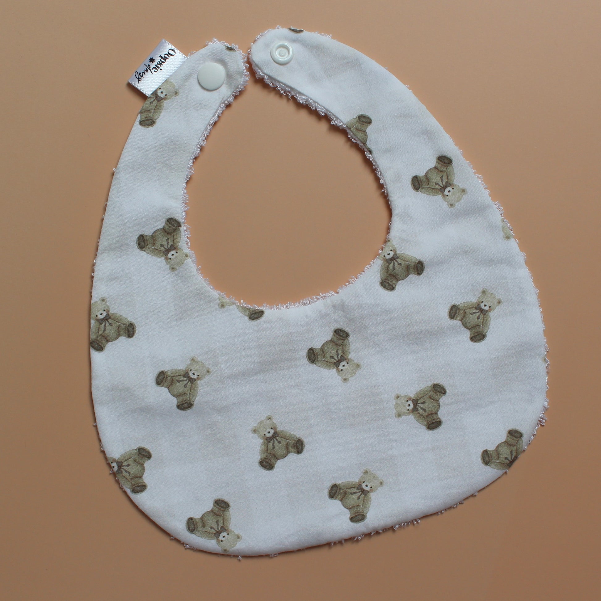 A white fabric baby bib with a repeating pattern of small brown teddy bears and subtle gingham pattern, featuring white snap closures at the top, laid flat on a solid peach-colored background.