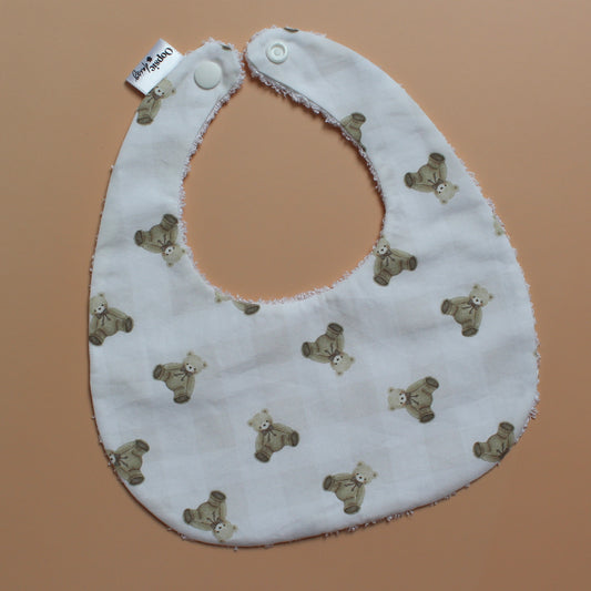 A white fabric baby bib with a repeating pattern of small brown teddy bears and subtle gingham pattern, featuring white snap closures at the top, laid flat on a solid peach-colored background.