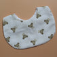 A white fabric bib with an all-over print of small brown teddy bears and subtle gingham pattern, shown on a solid tan background.