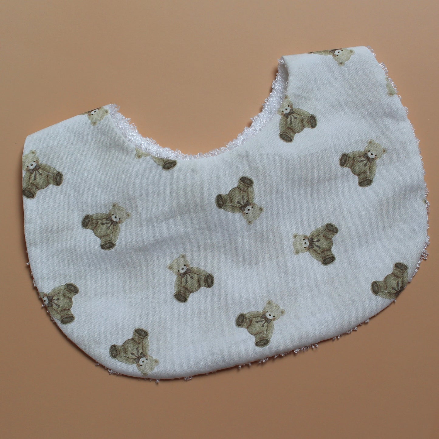 A white fabric bib with an all-over print of small brown teddy bears and subtle gingham pattern, shown on a solid tan background.