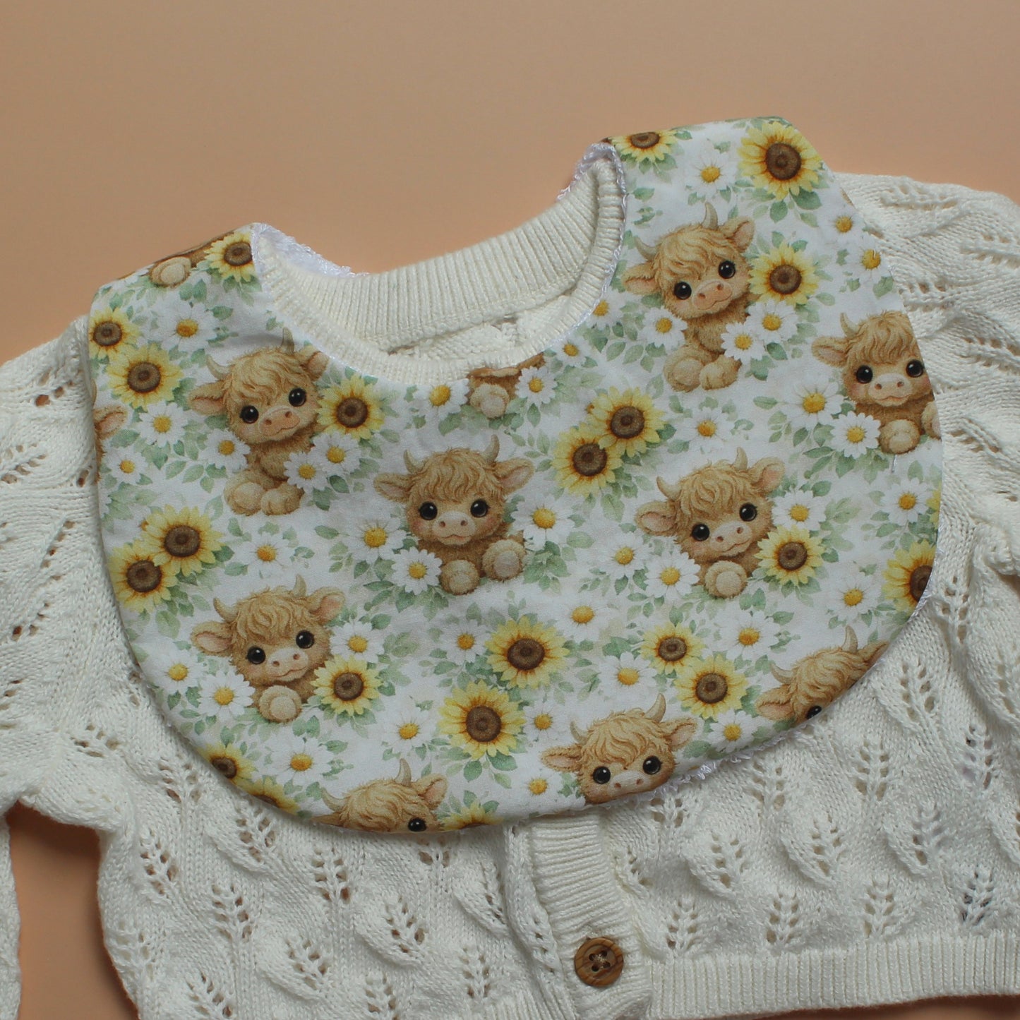 A close-up of a round baby  bib with a print of cute brown highland cow illustrations and yellow sunflowers, laid flat on a solid peach background.