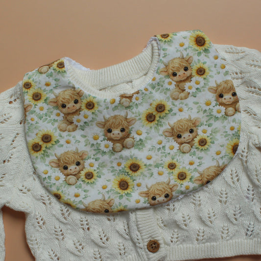 A close-up of a round baby  bib with a print of cute brown highland cow illustrations and yellow sunflowers, laid flat on a solid peach background.