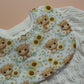 A close-up of a baby bib with a repeating pattern of Highland cows surrounded by yellow sunflowers with green leaves, layered over a cream-coloured cardigan