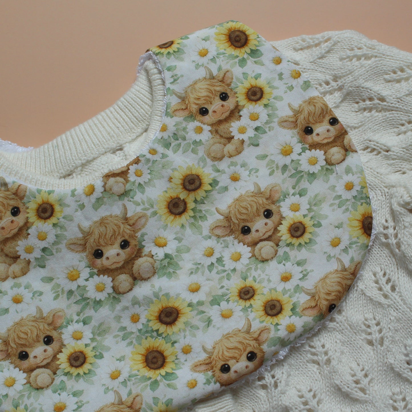 A close-up of a baby bib with a repeating pattern of Highland cows surrounded by yellow sunflowers with green leaves, layered over a cream-coloured cardigan