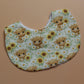 A baby bib with a white background printed with a pattern of cute yellow sunflowers and highland cows, laid flat on a solid tan background.