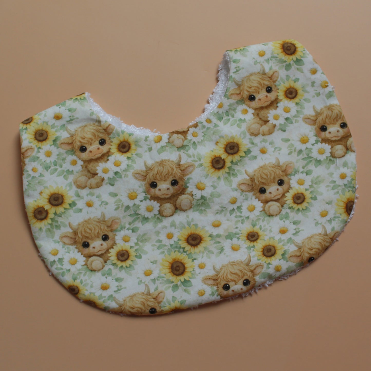 A baby bib with a white background printed with a pattern of cute yellow sunflowers and highland cows, laid flat on a solid tan background.