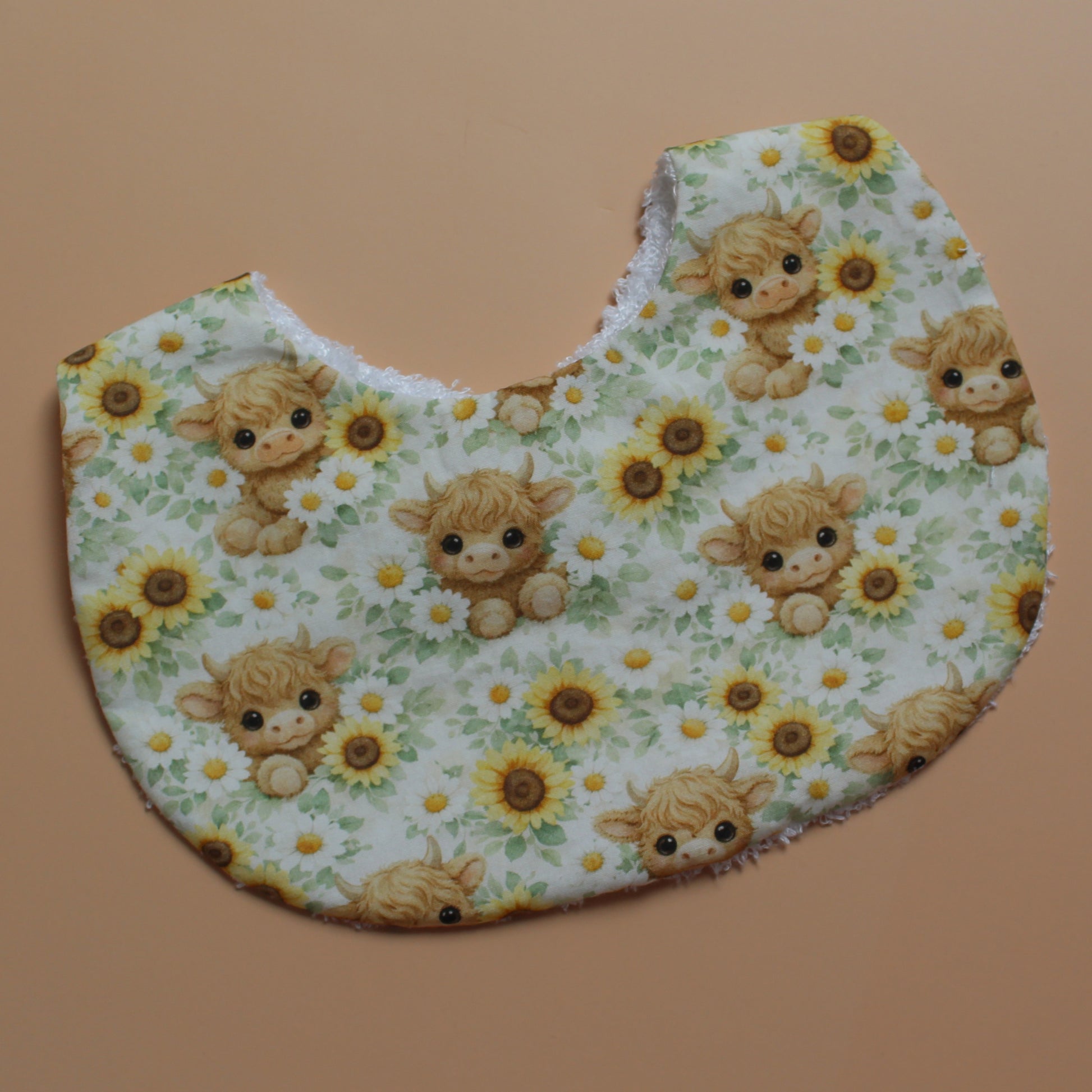 A baby bib with a white background printed with a pattern of cute yellow sunflowers and highland cows, laid flat on a solid tan background.
