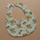 A baby bib with a highland cow and sunflower pattern, laid flat on a solid peach-coloured background. The bib has a white fabric base with a multicolour print of light brown highland cows, yellow sunflowers, and green leaves, and is secured with white snap buttons.