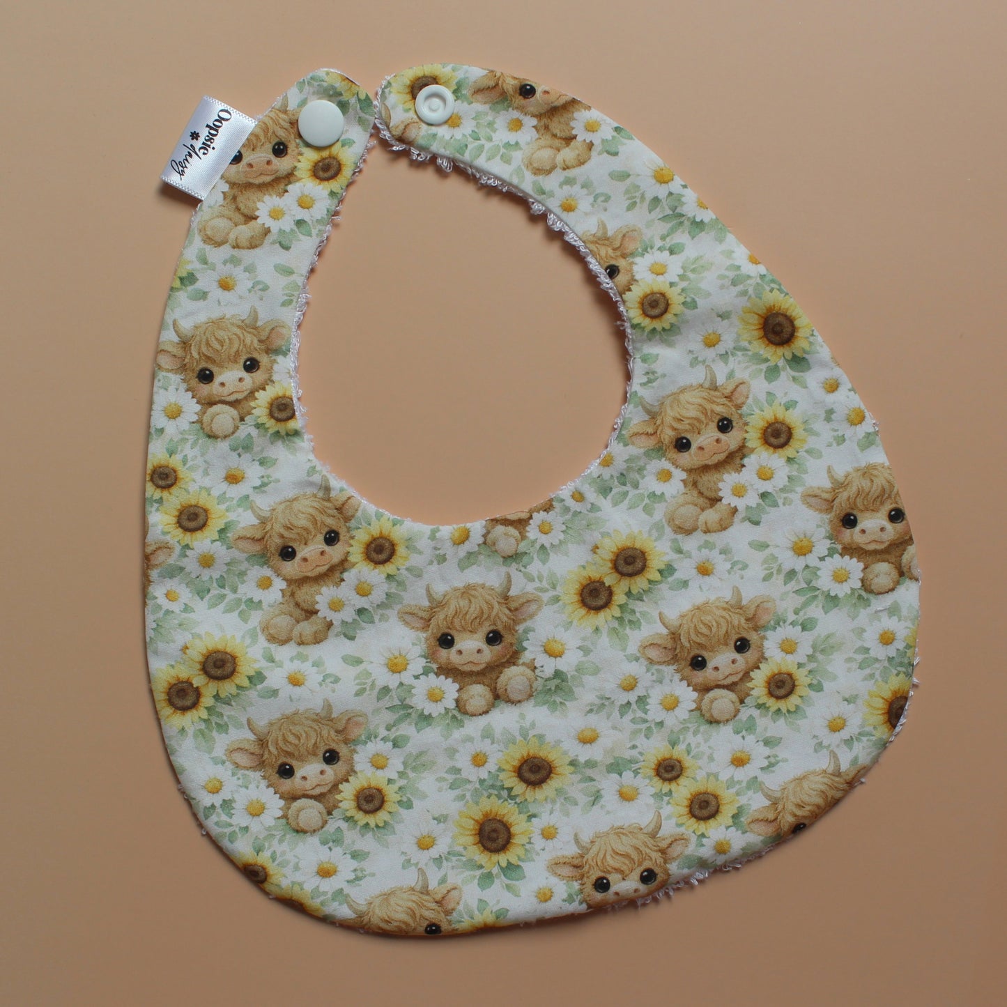 A baby bib with a highland cow and sunflower pattern, laid flat on a solid peach-coloured background. The bib has a white fabric base with a multicolour print of light brown highland cows, yellow sunflowers, and green leaves, and is secured with white snap buttons.