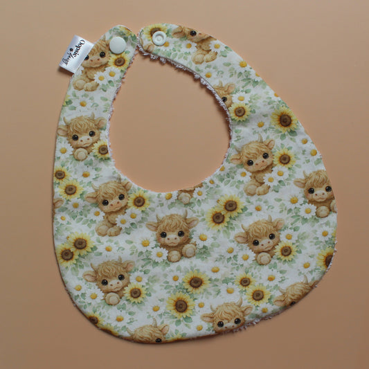 A baby bib with a highland cow and sunflower pattern, laid flat on a solid peach-coloured background. The bib has a white fabric base with a multicolour print of light brown highland cows, yellow sunflowers, and green leaves, and is secured with white snap buttons.