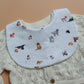 A white baby bib with a colourful print of various dog illustrations, styled over a cream-coloured knit cardigan.