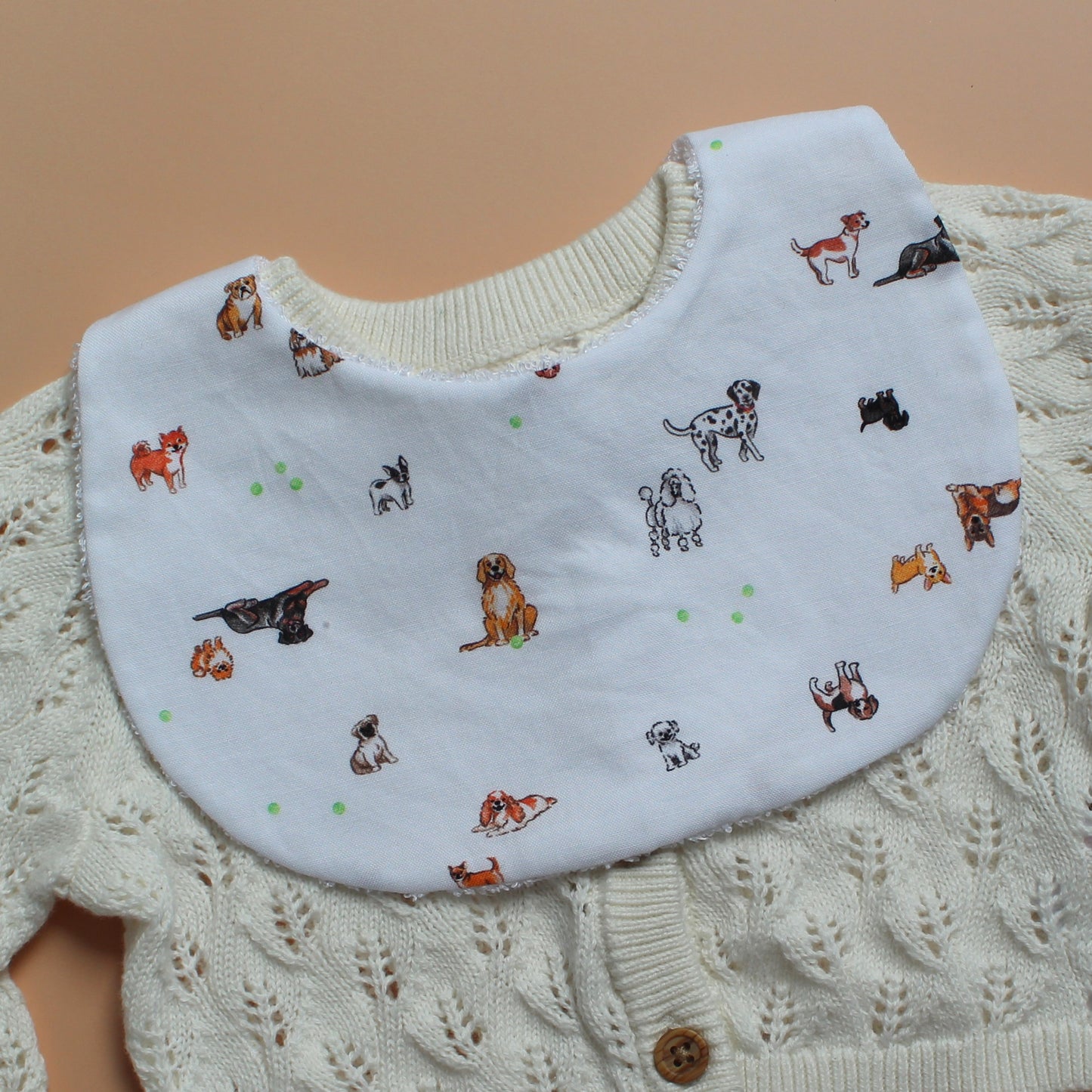 A white baby bib with a colourful print of various dog illustrations, styled over a cream-coloured knit cardigan.
