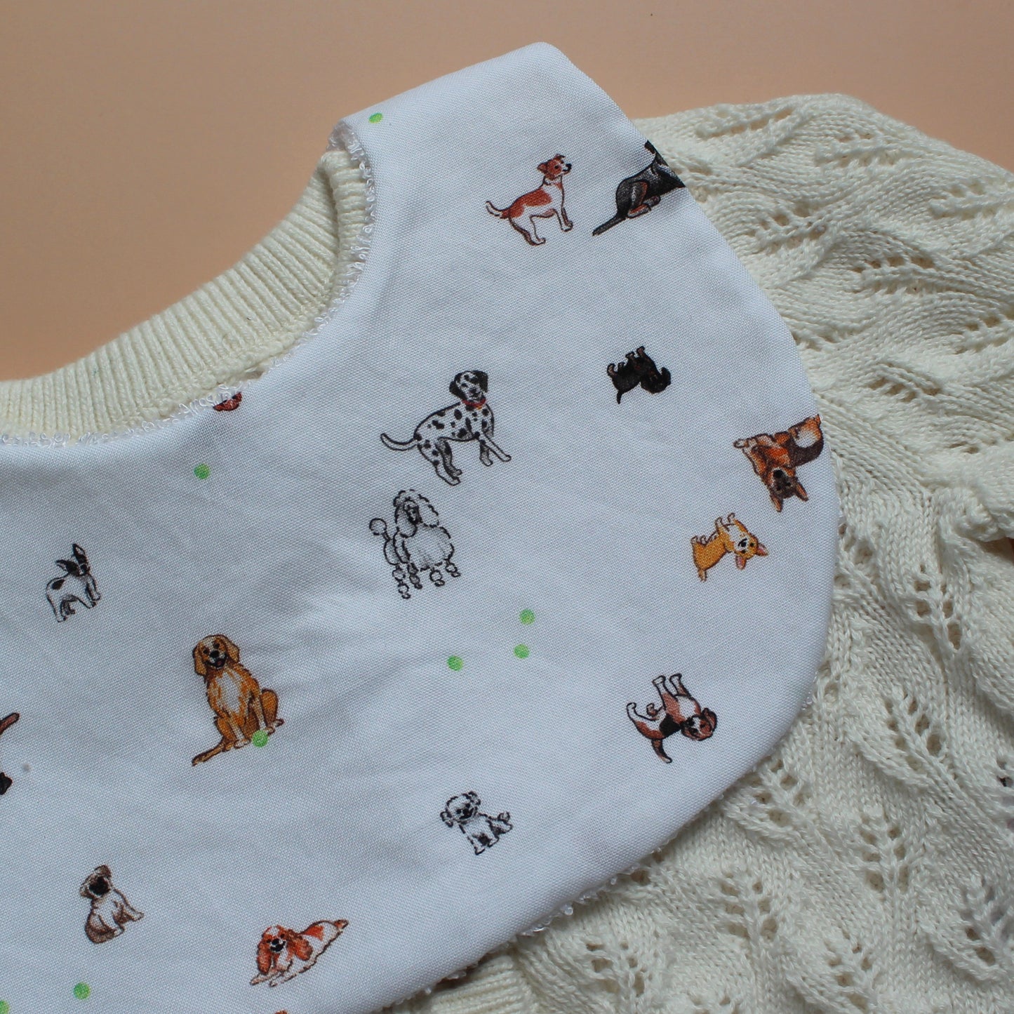 A close-up of a white baby bib with a dog print, laid over a cream-coloured cable-knit cardigan on a solid tan background.