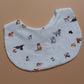 A white fabric bib printed with various dog illustrations in black, brown, and white, accented with small green tennis balls, laid flat on a solid tan background.