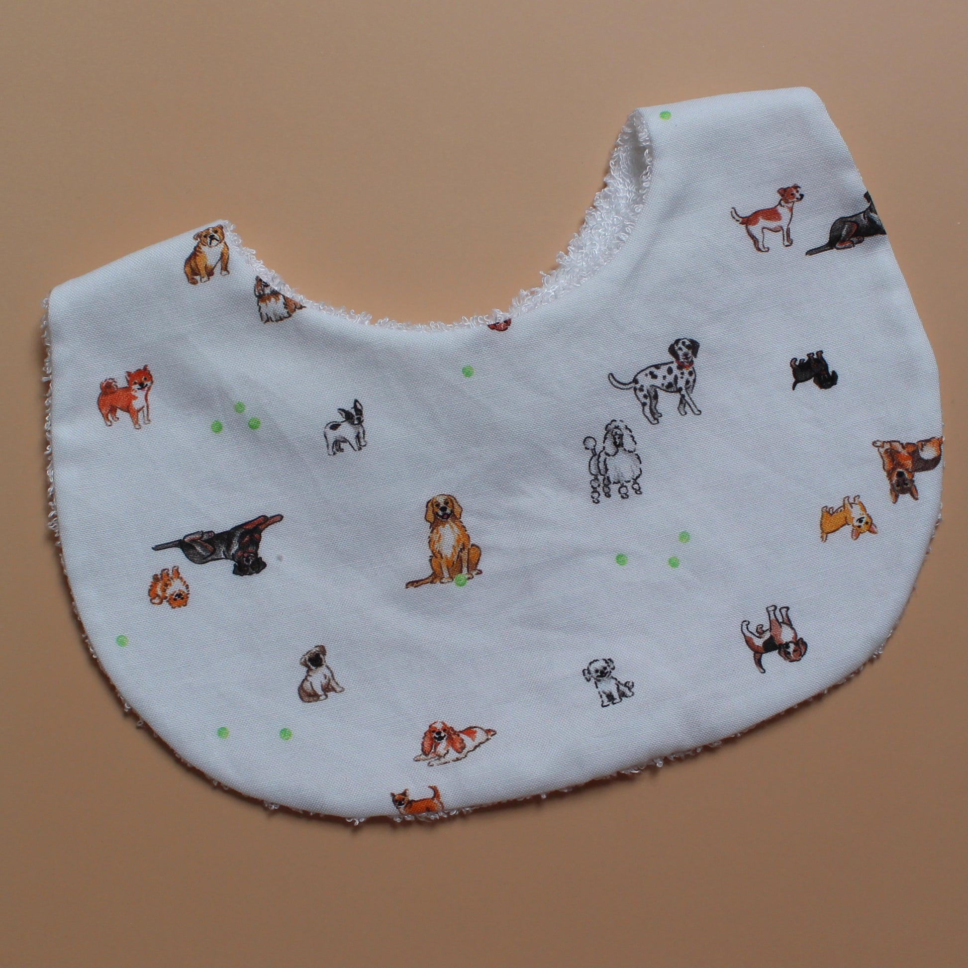 A white fabric bib printed with various dog illustrations in black, brown, and white, accented with small green tennis balls, laid flat on a solid tan background.