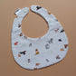 A white fabric bib with a playful print of various dog breeds in brown, black, and white, accented with green tennis balls. It has white snap buttons at the top and a small brand tag on the left side, photographed flat on a solid tan background.