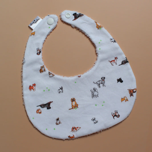 A white fabric bib with a playful print of various dog breeds in brown, black, and white, accented with green tennis balls. It has white snap buttons at the top and a small brand tag on the left side, photographed flat on a solid tan background.
